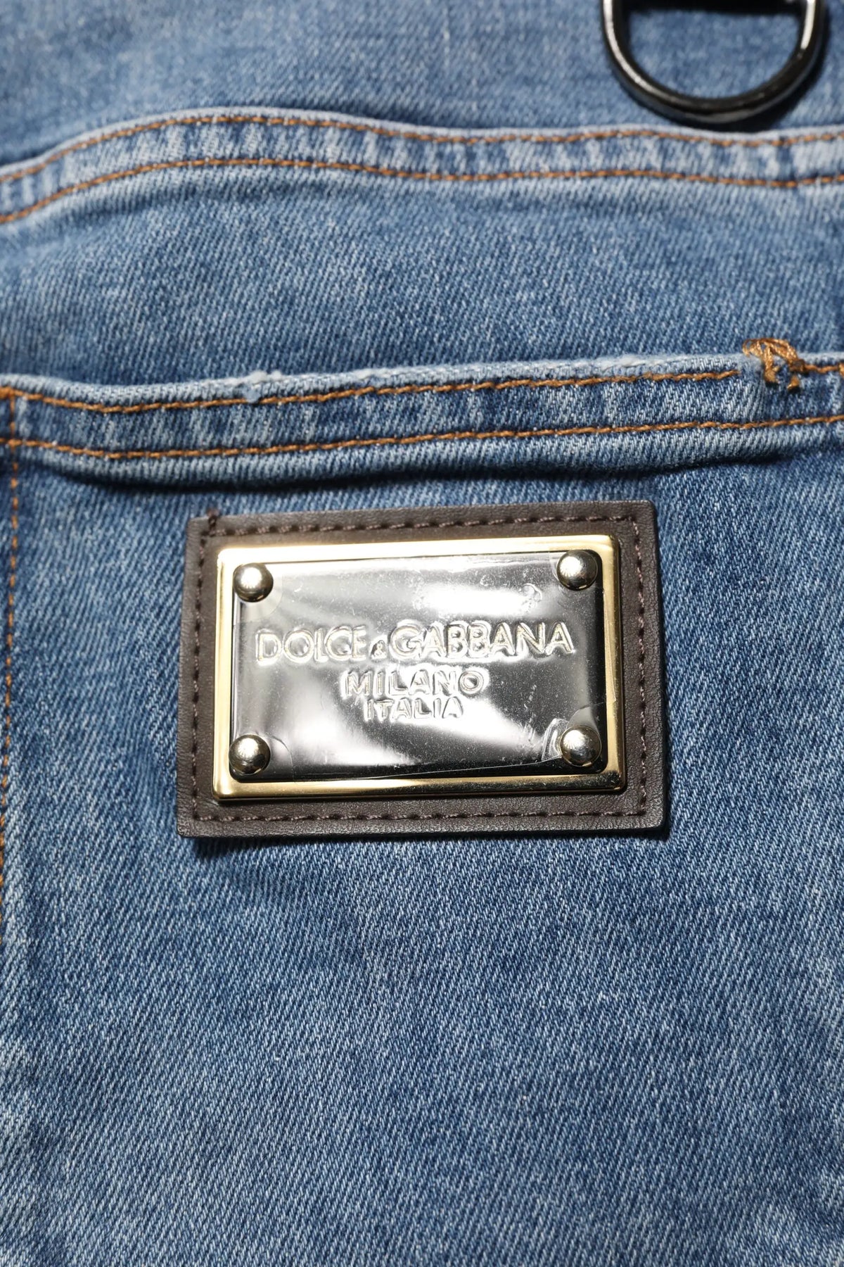 Plaque rectangulaire métallique dolce & gabbana milano italia encadrée de cuir marron foncé sur jean denim skinny bleu en coton déchiré