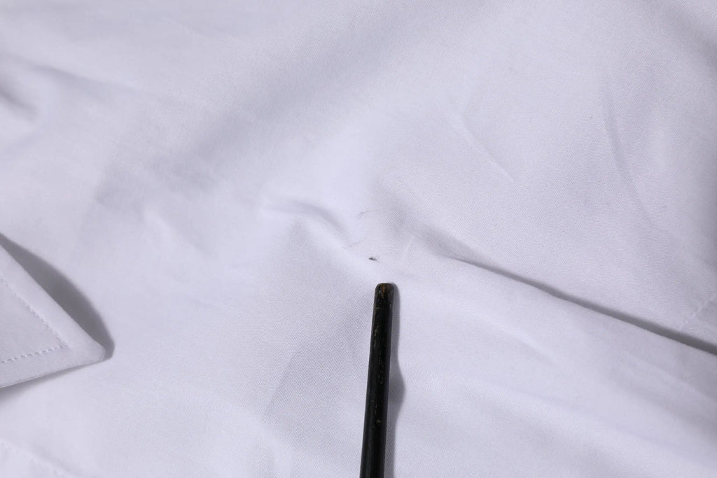 Un crayon noir mince posé en diagonale sur un tissu blanc à côté d’un chemisier blanc en coton à manches longues dolce & gabbana