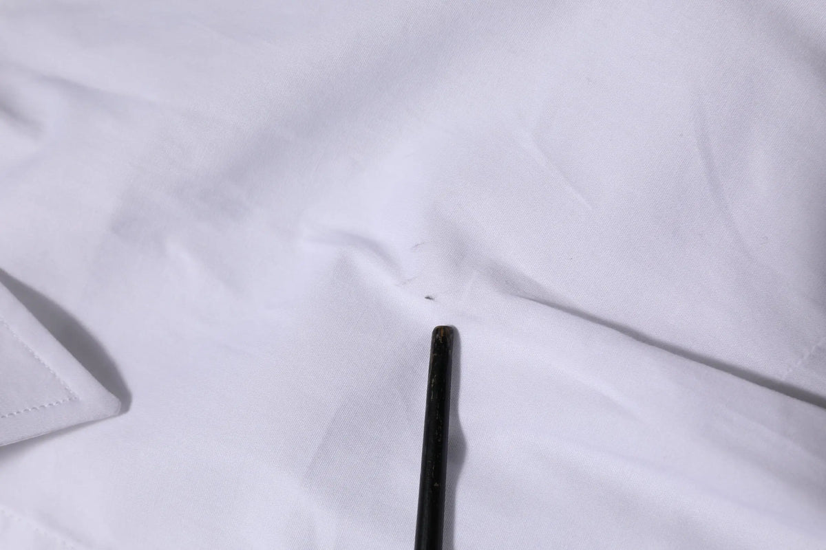 Un crayon noir mince posé en diagonale sur un tissu blanc à côté d’un chemisier blanc en coton à manches longues dolce & gabbana
