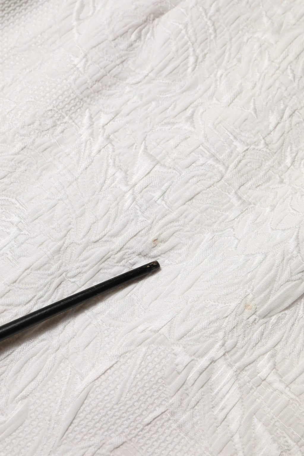 Objet cylindrique noir mat couché en diagonale sur tissu blanc texturé avec jupe dolce & gabbana blanche en brocart floral