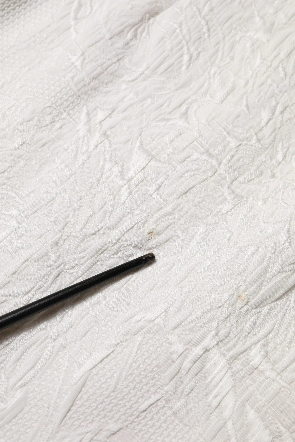 Objet cylindrique noir mat couché en diagonale sur tissu blanc texturé avec jupe dolce & gabbana blanche en brocart floral