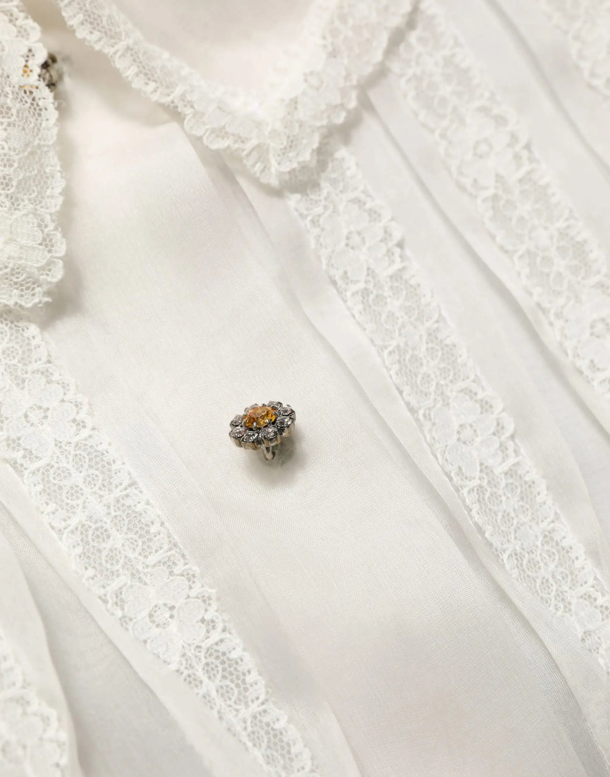 Broche délicate en forme de fleur avec pierre dambre centrale et petites pierres claires en métal argenté sur blouse blanche en soie transparente à volants dolce gabbana